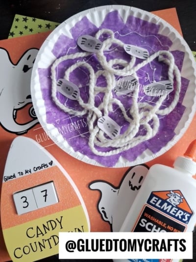 A Halloween craft setup with ghost cutouts, a candy countdown card displaying 37, a bottle of Elmers glue, and a paper plate spider web craft made from yarn and paper. @GLUEDTOMYCRAFTS is labeled at the bottom.