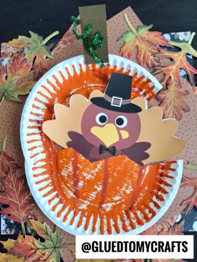 A Thanksgiving craft featuring a paper plate painted orange like a pumpkin, decorated with a cartoon turkey wearing a pilgrim hat and bow tie. Perfect for turkey kid crafts, with autumn leaves and the handle @GLUEDTOMYCRAFTS visible.