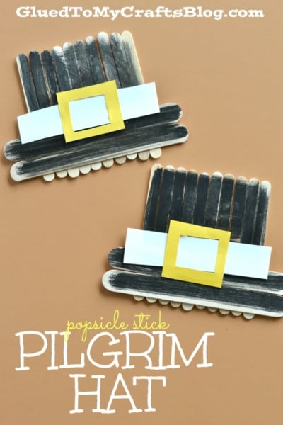 Two handmade craft stick pilgrim hats, created from black popsicle sticks, white paper bands, and yellow paper buckles, are displayed on a light brown surface. Text reads Popsicle Stick Pilgrim Hat—a fun pilgrim hat craft idea for kids.