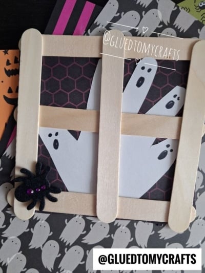 Paper ghosts and a spider are framed by popsicle sticks, resembling a charming Popsicle Stick Ghost Window. The background is decorated with ghost and bat patterns. @GLUEDTOMYCRAFTS is displayed on the image.