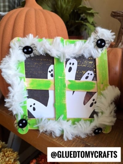 A green popsicle stick picture frame, designed as a Popsicle Stick Ghost Window, is decorated with fluffy white trim and black spider shapes surrounding ghost cutouts, with pumpkins in the background. Text @GLUEDTOMYCRAFTS appears at the bottom.