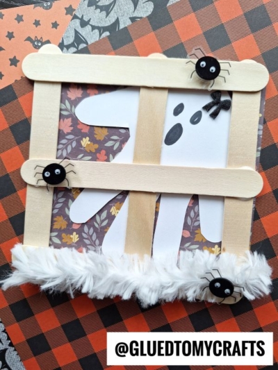 A cute Popsicle Stick Ghost Window craft with sticks forming a window over a ghost drawing, decorated with small spiders and fluffy material. A bat and Halloween-patterned paper complete this fun Halloween decoration.
