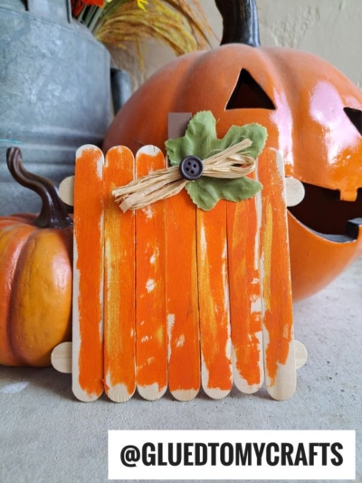 A square popsicle stick pumpkin, made from orange-painted sticks and decorated with raffia, a green leaf, and a button, sits in front of two pumpkins. @GLUEDTOMYCRAFTS is written at the bottom. Perfect for Popsicle Stick Pumpkins fun!.