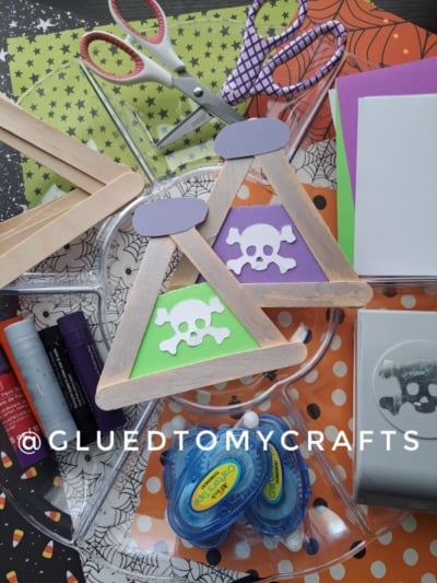 Craft supplies, including scissors, markers, glue, foam sheets, a Popsicle Stick Beaker craft, and two triangle popsicle stick crafts with skull and crossbones cutouts, are arranged on a Halloween-themed background. @GLUEDTOMYCRAFTS is written across the image.