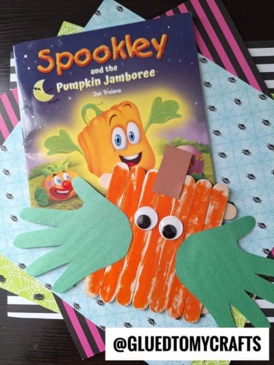 A children’s book titled *Spookley and the Pumpkin Jamboree* with a popsicle stick pumpkin craft made of orange popsicle sticks, green paper hands, and a brown stem, displayed on colorful paper backgrounds.