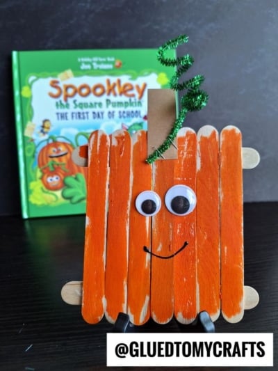 A charming popsicle stick pumpkin craft made from orange popsicle sticks with googly eyes, a smile, a clothespin stem, and a green pipe cleaner sits in front of a “Spookley the Square Pumpkin” book. @GLUEDTOMYCRAFTS text is at the bottom—a fun way to create your own popsicle stick pumpkins!.