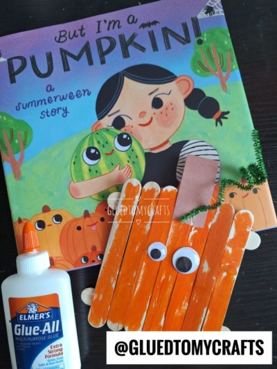 Children’s book “But I’m a Pumpkin!” with a girl hugging a pumpkin, next to a popsicle stick pumpkin craft made from orange sticks, googly eyes, green pipe cleaner, brown paper, and Elmer’s Glue.