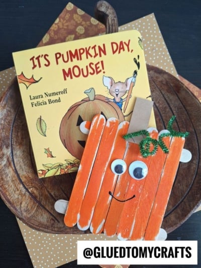 A children’s book titled “It’s Pumpkin Day, Mouse!” sits next to a popsicle stick pumpkin craft made from orange-painted sticks with googly eyes, a smile, and a green pipe cleaner, on a wooden plate. Text: @GLUEDTOMYCRAFTS.