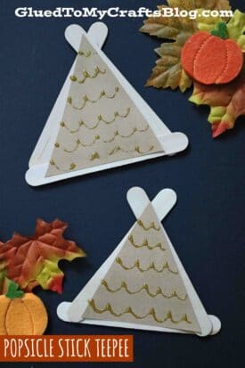 Two craft stick teepees decorated with gold wavy lines on tan paper, surrounded by artificial autumn leaves and small pumpkins, displayed on a dark background.