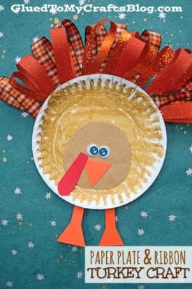 A Paper Plate & Ribbon Turkey craft made from a brown painted paper plate, with googly eyes, an orange beak, red snood, orange legs, and red plaid ribbon feathers, displayed on a blue background with small white stars.