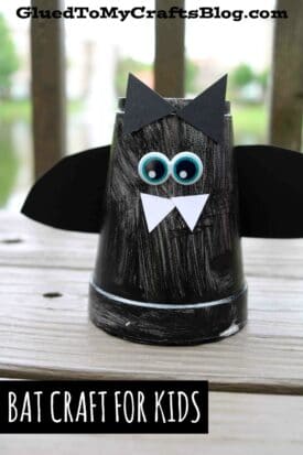 A black-painted cup bat, decorated with paper ears, wings, googly eyes, and white fangs, sits on a wooden surface. Text reads “Bat Craft for Kids” and “GluedToMyCraftsBlog.com.”.