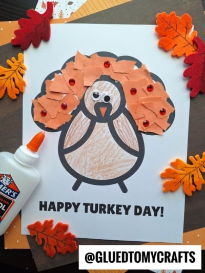 A turkey coloring page is decorated with orange torn paper and googly eyes. Red beads and felt leaves surround the page, along with Elmers glue—perfect inspiration for creative turkey kid crafts! The text reads HAPPY TURKEY DAY! and @GLUEDTOMYCRAFTS.