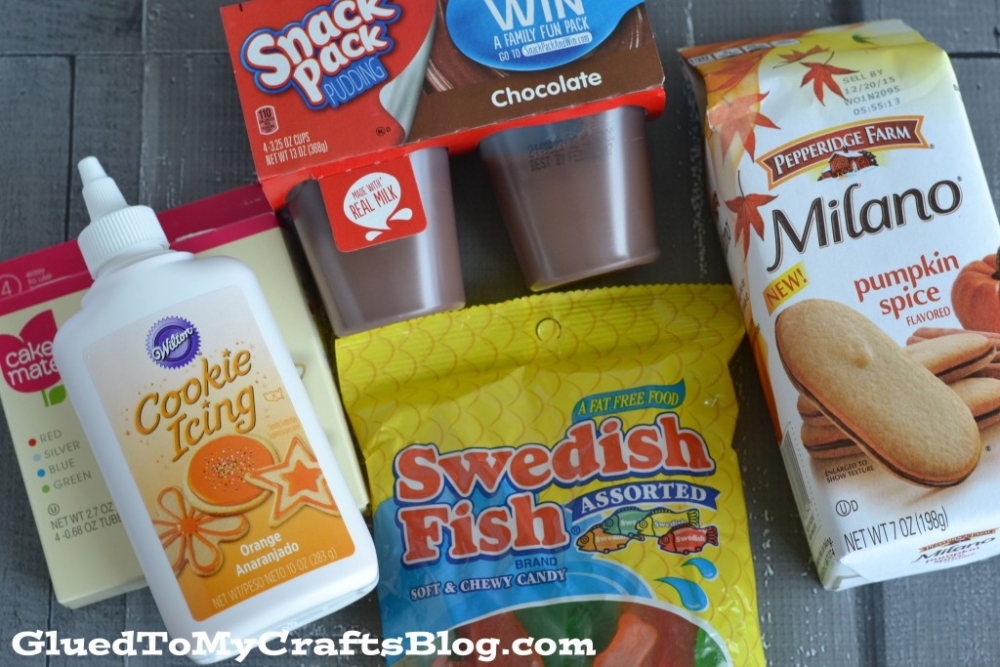 Assorted snacks and baking items on a table, including chocolate pudding cups, Milano pumpkin spice cookies, Swedish Fish candy, orange cookie icing, turkey pudding snacks, and a box of cake mix.