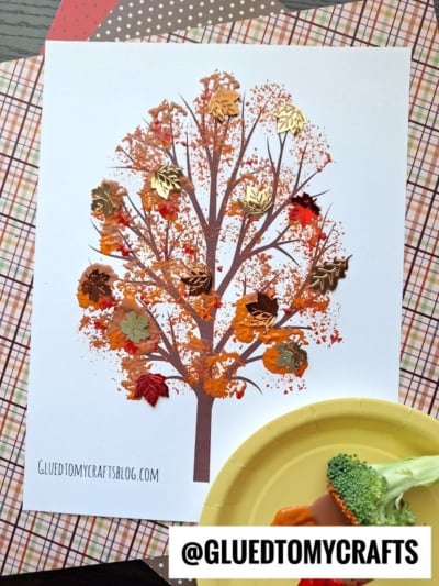 A broccoli stamped fall tree craft made with orange and brown paint, adorned with shiny metallic leaf stickers. A yellow plate with broccoli and a paintbrush sits nearby, all arranged on patterned paper.