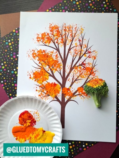 A broccoli stamped fall tree is created by painting a printed tree outline with autumn colors, using a broccoli floret as a stamp. Paint on a plate and colorful star paper are visible. Instagram handle @GLUEDTOMYCRAFTS is seen at the bottom.