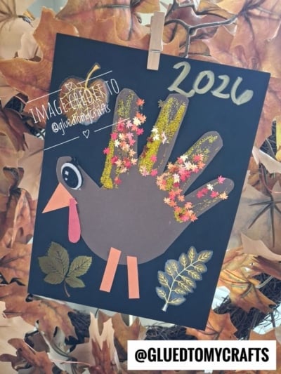 A handprint turkey craft on black paper, decorated with faux fall leaves and the year 2026, perfect for fall craft ideas.