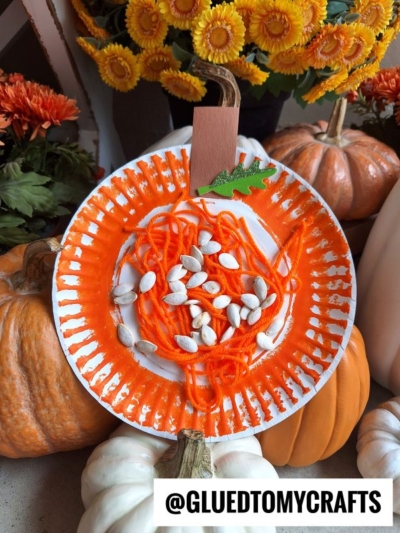A charming paper plate yarn pumpkin craft featuring orange yarn and pumpkin seeds, surrounded by real pumpkins and vibrant orange flowers. The Instagram handle @GLUEDTOMYCRAFTS is displayed at the bottom, making this yarn pumpkin kid craft a perfect seasonal activity.
