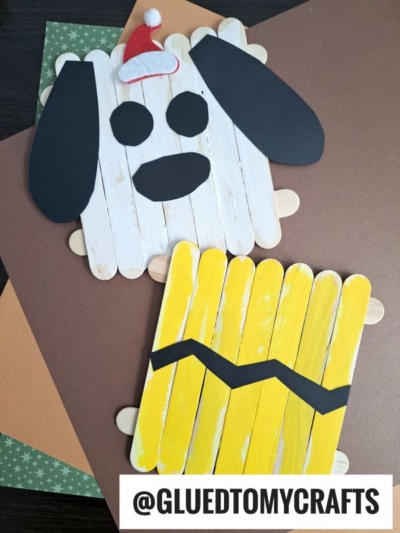 Two crafts made from popsicle sticks: one is painted white with black paper ears, eyes, nose, and a red Santa hat to resemble a dog; the other is a popsicle stick Charlie Brown with yellow paint and a black zigzag stripe like his shirt.