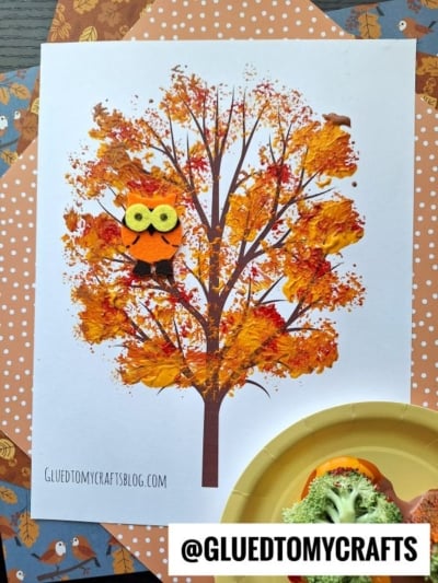 A broccoli stamped fall tree craft made with orange and yellow paint for leaves, featuring a small felt owl on a printed tree trunk, placed on polka dot paper with autumn-themed scrapbook paper in the background.