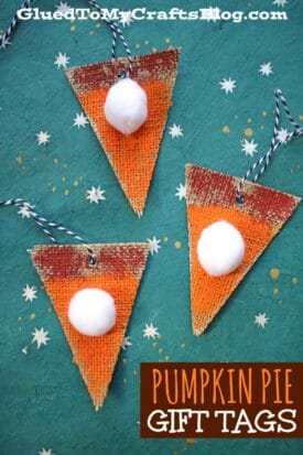 Three triangular Burlap Pumpkin Pie gift tags, painted orange with a white pom-pom on each, displayed on a teal background with white star patterns. Black and white string is attached for hanging.