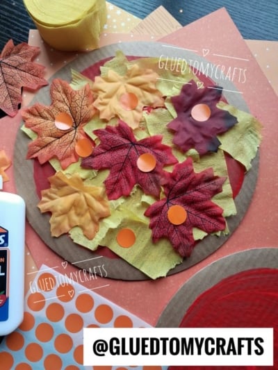 A fall-themed craft with faux leaves, orange dot stickers, yellow tissue paper, and a glue bottle on a round paper base—perfect for anyone searching for creative Fall Craft Ideas. Displayed on a table with @GLUEDTOMYCRAFTS.