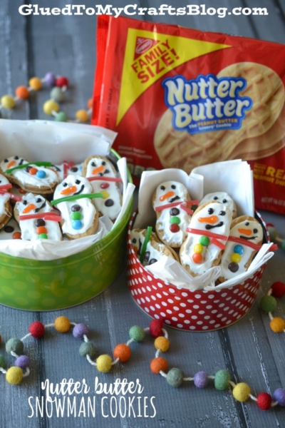 Two festive tins are filled with nutter butter snowmen cookies, each decorated as cheerful snowmen with icing and candy. A Family Size Nutter Butter package sits in the background, surrounded by playful pom-poms on a wooden surface.
