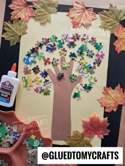 A paper hand forms a tree trunk on yellow paper, with puzzle pieces as leaves—a creative pick for Fall Craft Ideas. Bottled glue, tray, and faux autumn leaves surround the craft. @GLUEDTOMYCRAFTS is on the image.