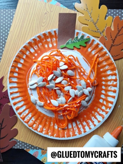 A paper plate yarn pumpkin with pumpkin seeds arranged on top to resemble pumpkin guts. Paper leaves, a brown paper rectangle, and a bottle of glue are nearby. The username @GLUEDTOMYCRAFTS appears at the bottom of this yarn pumpkin kid craft.