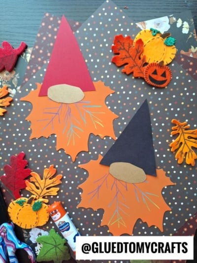 Two fall gnome crafts made with orange leaf shapes, colored paper hats, and paper noses are displayed on a polka dot background with felt leaves, pumpkins, a glue stick, and the text @GLUEDTOMYCRAFTS—perfect for anyone looking for fun Fall Craft Ideas.