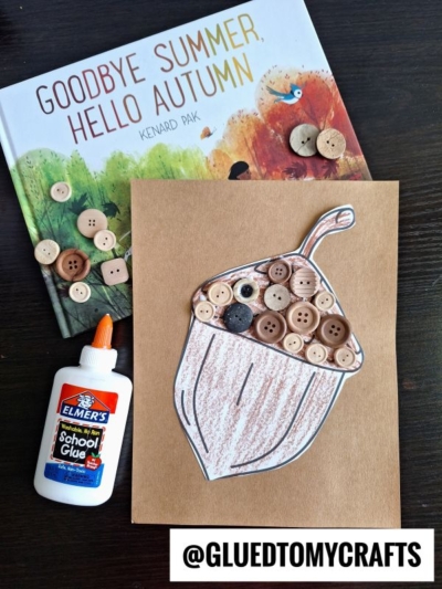A children's book titled Goodbye Summer, Hello Autumn, a bottle of Elmer's glue, and a button acorn craft decorated with brown buttons are arranged on a dark wooden surface. Text reads @GLUEDTOMYCRAFTS.