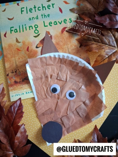A children’s book titled Fletcher and the Falling Leaves is shown with a fox craft made from brown paper, googly eyes, and a round nose, surrounded by artificial autumn leaves—perfect inspiration for fun fall craft ideas.
