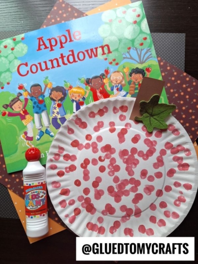 A children’s book titled Apple Countdown, a polka-dotted paper plate apple craft, green leaf, brown stem, red dot marker, and colored paper make for fun Fall Craft Ideas, all arranged on a dark surface.