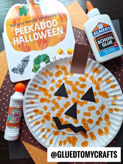 A paper plate decorated as a pumpkin with orange dot paint and black paper cutouts for the face sits next to glue, dot markers, and the book The Very Hungry Caterpillar’s Peekaboo Halloween—a perfect addition to your Fall Craft Ideas.