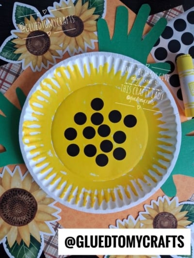 A paper plate painted yellow with black dots to resemble a sunflower, surrounded by sunflower cutouts, green paper leaves, and a glue stick. The plate is set on a colorful background, making it a cheerful addition to your collection of Fall Craft Ideas.