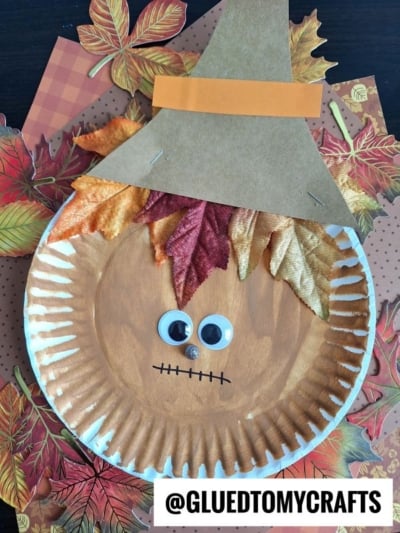 A Paper Plate Scarecrow craft with a brown painted face, googly eyes, faux autumn leaves for hair, a brown paper hat, and a stitched smile sits on colorful fall leaves. @GLUEDTOMYCRAFTS is written at the bottom.