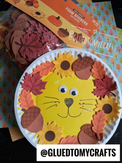 A paper plate lion face craft with googly eyes, made with yellow paint and decorated with foam leaves, acorns, and sunflowers is the perfect addition to your collection of Fall Craft Ideas. A bag of foam stickers and patterned paper are in the background.