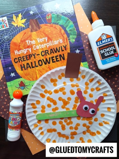 A Halloween-themed craft setup featuring The Very Hungry Caterpillar’s Creepy Crawly Halloween book, Elmer’s glue, a bingo dauber, and a paper plate caterpillar craft with orange dots and googly eyes—perfect for fall craft ideas.