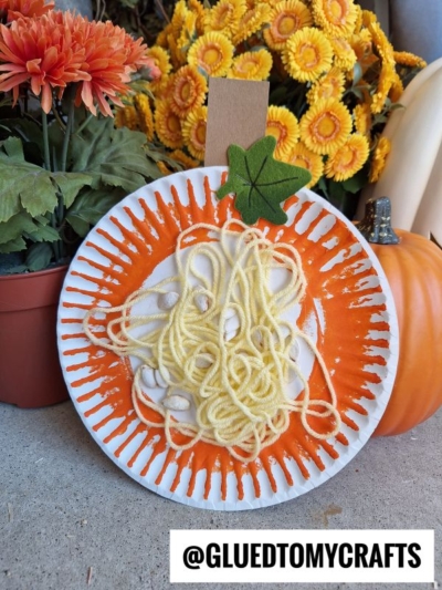 A paper plate yarn pumpkin kid craft with orange paint, yellow yarn, and a green paper leaf sits near orange and yellow flowers and a pumpkin. @GLUEDTOMYCRAFTS is written at the bottom.