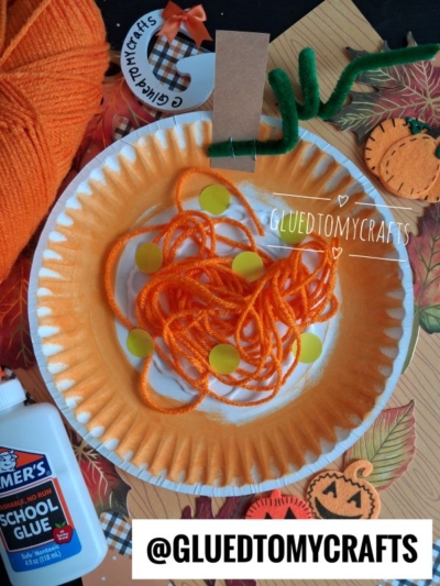 A paper plate yarn pumpkin painted orange, with orange yarn and yellow dots glued on, perfectly resembling a festive fall pumpkin. Surrounded by craft supplies—yarn, glue, pipe cleaner, paper cutouts—and a @GLUEDTOMYCRAFTS sign. This yarn pumpkin kid craft is a fun and creative activity for little hands!.