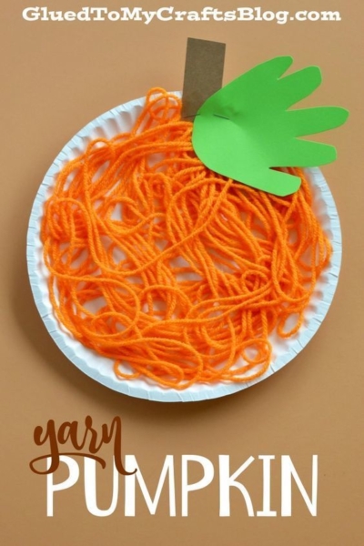 Paper plate with orange yarn arranged like a pumpkin, a green paper leaf shaped like a hand, and a brown paper stem on top, against a brown background. This charming yarn pumpkin kid craft features “Yarn Pumpkin” written at the bottom.