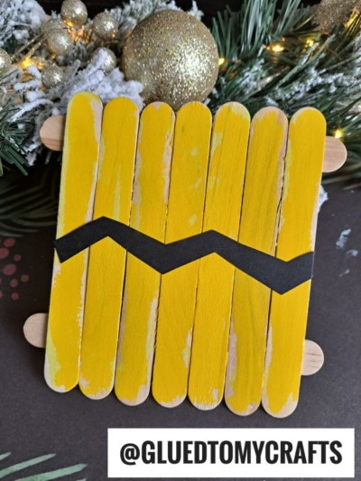 A popsicle stick Charlie Brown craft made of yellow-painted sticks with a black zigzag stripe, resembling his shirt. Pine branches, a gold ornament, and snow add festive charm. Text: @GLUEDTOMYCRAFTS.