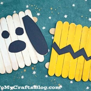 Two popsicle stick crafts on a blue surface with white stars: one is Snoopy’s face, and the other features a popsicle stick Charlie Brown yellow shirt with his signature black zigzag pattern.