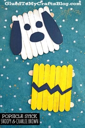 Two popsicle stick crafts on a blue surface with white stars: one is Snoopy’s face, and the other features a popsicle stick Charlie Brown yellow shirt with his signature black zigzag pattern.