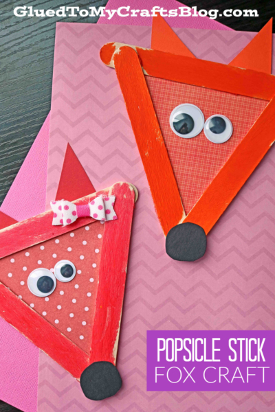 Two craft stick fox faces made from orange popsicle sticks, googly eyes, and black paper circles, arranged on pink and purple zigzag paper. One fox has a pink polka-dot bow. Text reads: POPSICLE STICK FOX CRAFT.