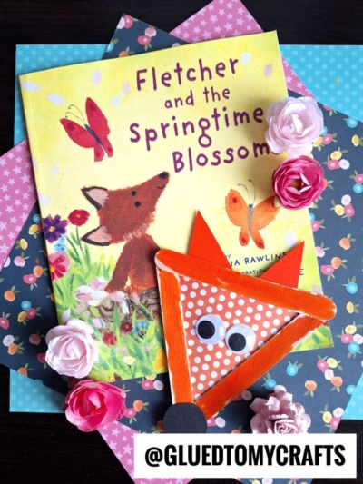A children's book titled Fletcher and the Springtime Blossom is surrounded by paper flowers and colorful polka-dotted paper, with a popsicle stick fox bookmark featuring googly eyes in the foreground.