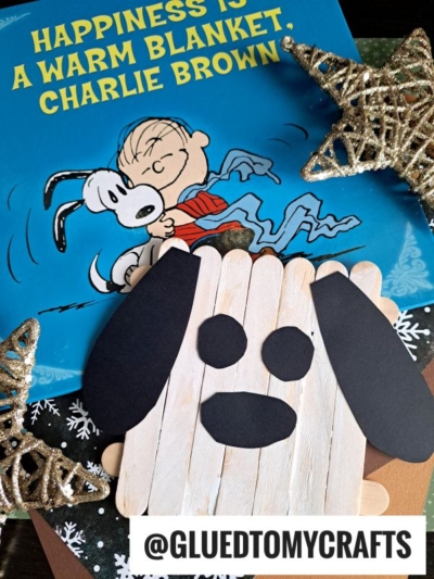 A Peanuts book titled Happiness Is a Warm Blanket, Charlie Brown lies beneath a handmade popsicle stick Charlie Brown craft, with glittery gold star decorations scattered around them.