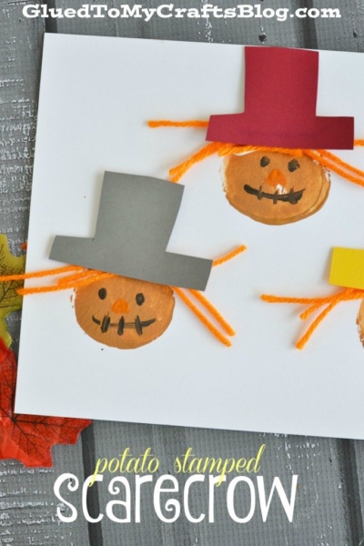 Three scarecrow faces stamped with potatoes are decorated with paper hats and orange yarn hair on white paper. The phrase “potato stamped scarecrow” is written at the bottom, and autumn leaves accent the left side, adding a festive seasonal touch.