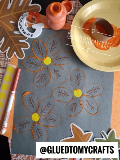 A craft setup with a black paper featuring orange and yellow flower drawings, a paint bottle, a brush, brown and orange markers, autumn leaf cutouts, and the username @GLUEDTOMYCRAFTS appearing twice—perfect inspiration for your next Fall Craft Ideas project.