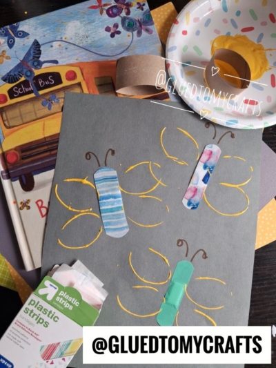 Childrens butterfly craft with bandages on gray paper, yellow paint for wings, art supplies, a book, and a paper plate with yellow paint—perfect for Fall Craft Ideas. Gluedtomycrafts watermark.