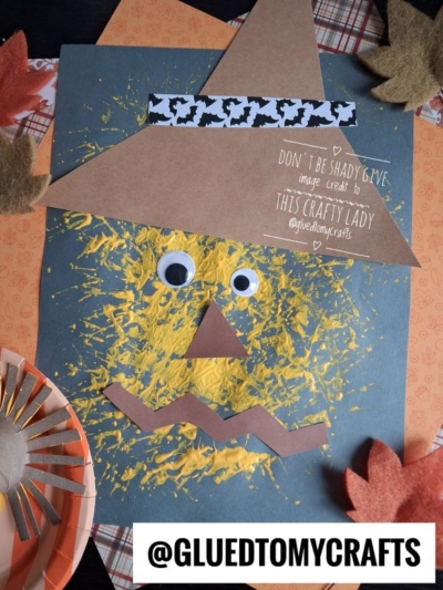 A scarecrow craft made with yellow yarn, googly eyes, a brown hat, and a zigzag paper mouth on blue paper. Surrounded by fake leaves and a paper plate, this fun project is perfect for anyone looking for creative Fall Craft Ideas. Text reads @GLUEDTOMYCRAFTS.
